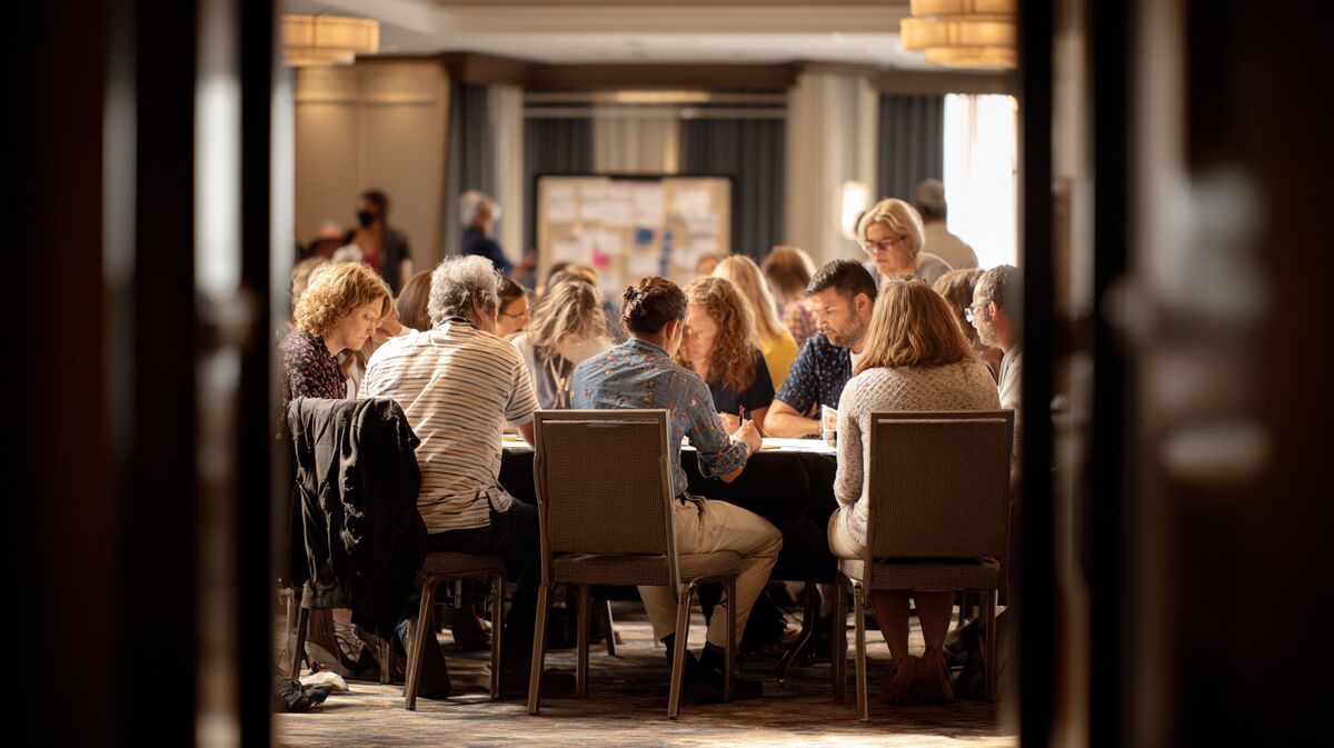 Coach faciliteert team workshop met diverse groep aan grote tafel, actieve participatie en samenwerking