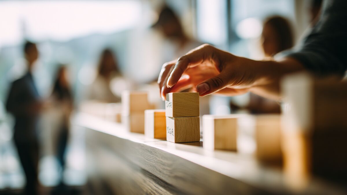 Coach arrangeert houten blokken met team development stages, workshop setting met deelnemers op achtergrond